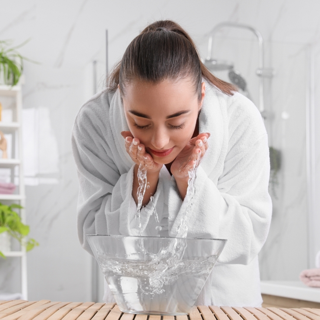 Woman in a white bathrobe washing her face with water from a large glass bowl in a bathroom after using MHOD's Facial Cleanser